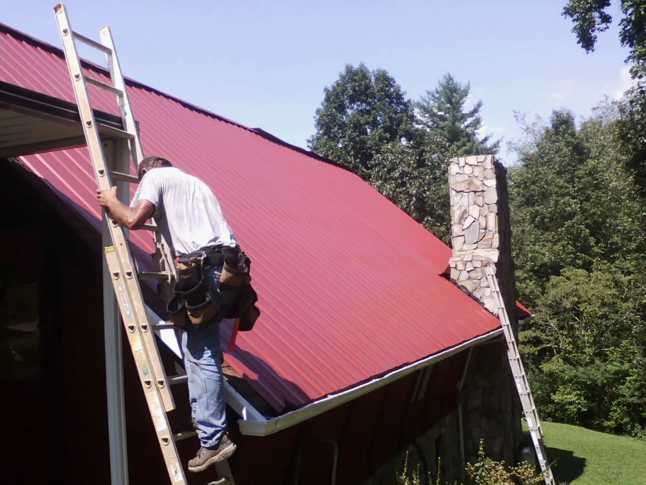 Roofer on a ladder beside a red metal roof and stone chimney — Soffit & Fascia Repair by Roofing Brothers in Fort Laramie
